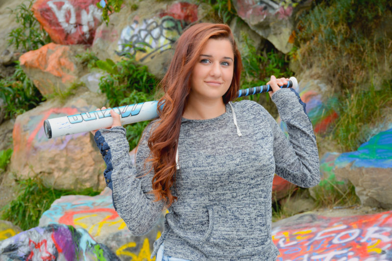 Senior girl posing with softball bat for senior portraits with a graffiti rock wall.